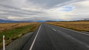 Bir araç, Queenstown, Yeni Zelanda yakınlarındaki açık tarım arazisi boyunca düz, boş kırsal bir yolda, bulutlu gökyüzünün altında, yumuşak doğal aydınlatma ve sabit ileri kamera hareketleriyle ilerliyor.