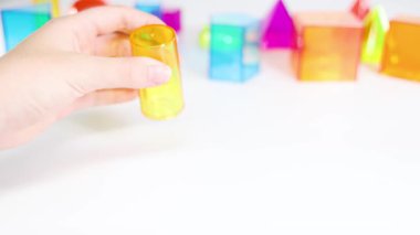 Eller, eğitici oyun ve dokunsal öğrenmeyi vurgulayarak, beyaz bir masa üzerindeki canlı üç boyutlu geometrik şekillerle etkileşir.