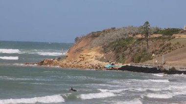 Güneşli bir günde bir sörfçü manzaralı bir uçurumun kenarında dalgalar sürer. Video, dinamik okyanus hareketini ve canlı kıyı manzarasını yakalıyor.