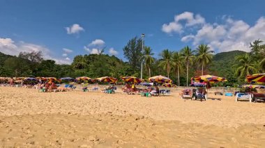 Turistler rahatlar ve Phuket, Tayland 'da güneşli bir plaj boyunca canlı şemsiyeler ve açık mavi bir gökyüzü ile yürürler.