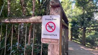 Parlak doğal gün ışığı altında, yemyeşil ormanlarla çevrili ahşap bir tahta kaldırımdaki 'Drone Free National Park' tabelasının önünden el kamerası geçiyor.