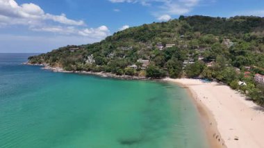 İnsansız hava aracı görüntülerinde turkuaz suları ve yemyeşil yemyeşil yemyeşil, parlak gün ışığında lüks villalar sergilenen huzurlu bir Phuket plajı görülüyor.