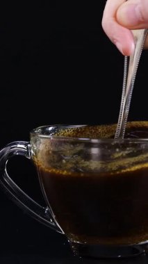 Bir el, temiz bir cam bardakta koyu bir fonda kahveyi karıştırır ve dönen bir hareket yaratır.