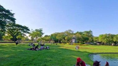 İnsanlar Bangkok 'un Rama IX Parkı' nda güneşli bir günün tadını çıkarıyorlar. Etrafı yeşillik ve dingin bir gölet ile çevrili. Canlı ve huzurlu bir atmosfer.