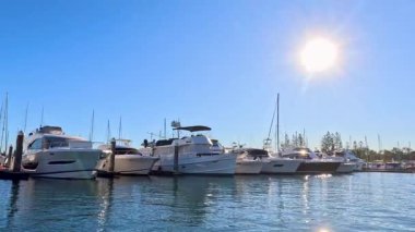 Lüks yatlar ve tekneler parlak güneş ışığı altındaki bir marinada demirli. Sakin sularda kameralar ve açık mavi gökyüzü var.