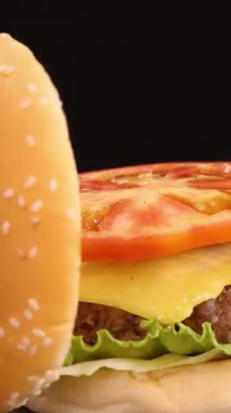 Susamlı çörek, salatalı, domatesli ve peynirli çizburgerin üzerine düşer. Stüdyo ışıklandırmalı siyah arka planda ağır çekimde çekilir.