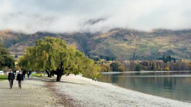 Wanaka Gölü 'nün kıyısında dağlar ve ağaçlarla çevrili bulutlu gökyüzünün altında yürüyen insanlar barışçıl ve manzaralı bir atmosfer yaratıyorlar.
