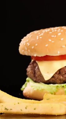 Altın patates kızartması ağır çekimde marul, domates ve peynirli bir çizburgerin yanına düşüyor. Parlak stüdyo ışıklarının altında siyah bir arkaplanda.