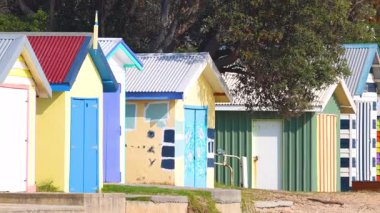 Canlı sahil kulübeleri, berrak gökyüzünün altındaki kumlu sahilde, sakin bir sahil ortamı sergiliyor.