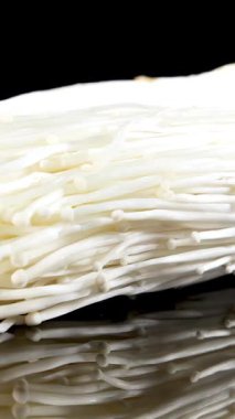 Bir parça enoki mantarı parlak stüdyo ışıkları altında parlak, siyah yansıtıcı bir yüzey üzerinde yavaşça döner, hassas dokularını ve kümelenmiş gövdelerini vurgular.