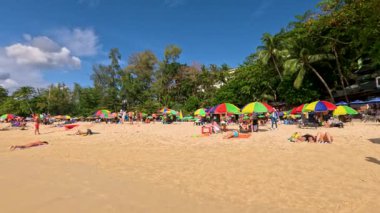 Phuket, Tayland 'da kum ve denizin tadını çıkaran insanların olduğu güneşli plaj sahnesi. Canlı şemsiyeler ve gür yeşillik tropikal bir atmosfer yaratır.