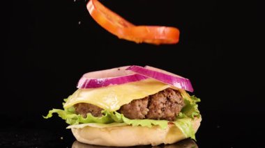 Susamlı çörek, salatalı, domatesli, soğanlı ve peynirli sığır etli burgerin üstüne düşer. Stüdyo ışıklandırmalı, siyah arka planda ağır çekimde çekilir.