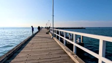 Bir insan güneşli bir iskelede yürür, berrak gökyüzü ve sakin bir deniz ile sakin bir atmosfer yaratır.