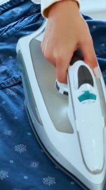 Bir insan buharlı ütüyle mavi desenli gömleğin kırışıklıklarını düzeltir. Yakın çekim, sabit kamera, ev dekorasyonu.