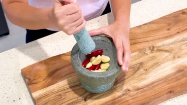 Kişi, sarımsak ve biber ezmek için taş havan ve tokmak kullanır. Parlak bir ahşap kesme tahtasının altında, hatta kamera açılı mutfak ışıklandırması bile.
