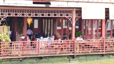 A vibrant riverside restaurant with patrons enjoying meals, captured from a moving boat on the Chao Phraya River