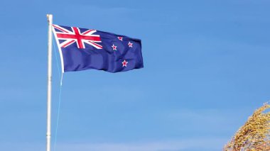 Yeni Zelanda bayrağı güçlü bir şekilde dalgalanır uzun bir bayrak direğinde berrak bir mavi gökyüzüne karşı, kayalık arazi görünür, parlak gün ışığında yakalanır