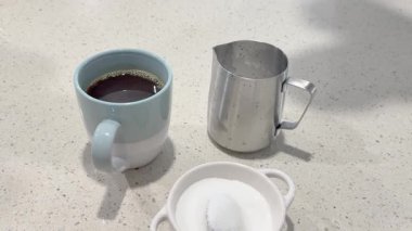 Bir el, mutfak tezgahındaki kahve bardağına şeker ekler, yanında da süt sürahisi.