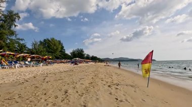 Turistler Phuket, Tayland 'da kumlu bir sahil boyunca yürüyorlar, parçalı bulutlu bir gökyüzünün altında, canlı plaj şemsiyeleriyle.