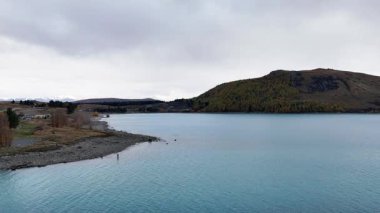 Tekapo Gölü 'nün sakin suları ve bulutlu gökyüzünün altındaki tepelerle sakin bir görüntüsü, sabit bir hava görüntüsüyle çekildi.