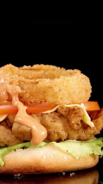 Salata, domates, peynir ve soğan halkalı kızarmış tavuk burgerinin yakın çekimi. Thousand Island sosu, dramatik stüdyo ışıkları altında dökülüyor.