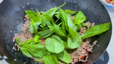 Taze fesleğen yaprakları tavada doğranmış ete ekleniyor ve parlak mutfak ışıkları altında kızartılıyor. Yakın çekim kamera açıları yemek pişirme sürecini yakalıyor.