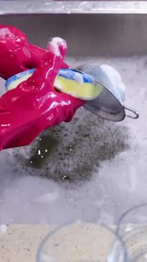 Pembe eldivenli eller sabunlu mutfak lavabosunda süngerle metal bir kevgir temizler.