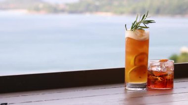 Bir el, deniz kenarındaki masadan bir kokteyli kaldırır ve okyanus manzaralı bir arka planda bir içki daha bırakır.