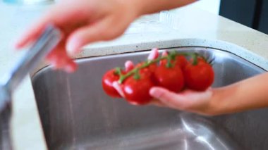 Eller, çağdaş bir mutfak lavabosunda akan suyun altında, parlak doğal ışıklandırma ve sabit kamera çerçeveli olgun sarmaşık domateslerini duruluyor.