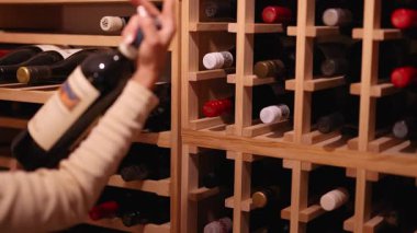 Bir kadın loş aydınlatılmış bir mahzende şarap şişelerini inceliyor, dikkatle ahşap bir raftan seçiyor.