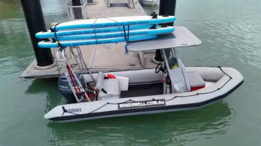 Drone, Port Douglas rıhtımında sörf tahtalarıyla şişme bot yakalıyor. Sakin sular, bulutlu ışıklar ve sabit kamera hareketleri.