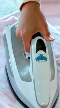 Bir kişi ütü masasına pembe bir gömlek basmak için modern buhar ütüsü kullanır, doğal gün ışığı ve kamera açıları ile süreci vurgular.