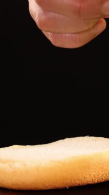 Bir el titizlikle taze bir marul yaprağını düz bir hamburger ekmeğine yerleştirir. Yakın plan, parlak stüdyo ışıklandırması ve siyah arka plan ile çekilir.