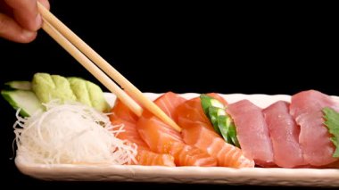 Yemek çubukları, stüdyo ışıklandırması altında düzgün düzenlenmiş bir tabaktan somon balığı sashimi ve istiridye suşisi parçaları seçer, taze deniz ürünleri ve canlı garnitürleri renklendirir.