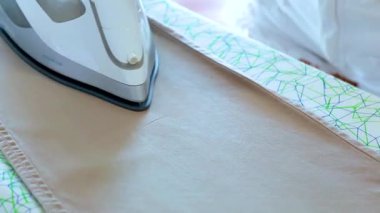 Bir buhar ütüsü desenli ütü masasında bej bir pantolonun üzerinden süzülür ve parlak, doğal gün ışığında buhar patlamaları yaratır. Kamera sabit duruyor, ütü sürecine odaklanıyor