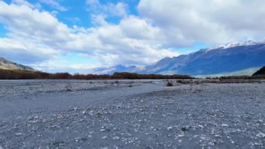 Parçalı bulutlu bir gökyüzünün altında uzak dağlarla geniş bir nehir yatağı. Yumuşak ışıklandırma ve minimum hareketle sakin, doğal ortam