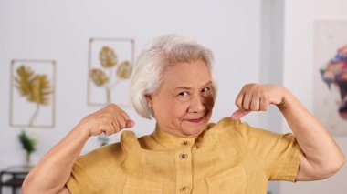 Sarı tişörtlü yaşlı Asyalı kadın kollarını açıp gülümsüyor. Güveni ve güveni ifade ediyor. Aydınlatılmış, modern bir iç mekan ve yumuşak gün ışığında.