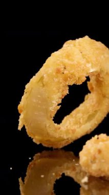 Altın kızartılmış soğan halkaları, parlak stüdyo ışıklarıyla aydınlanan parlak siyah bir yüzeye düşer ve yavaş çekimde dokuyu ve hareketi aydınlatır.