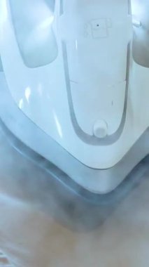 Bir buhar ütüsü desenli ütü masasında bej bir pantolonun üzerinden süzülerek görünür bir buhar yayar. Parlak, doğal ışıklandırma ve yakın çekim kamera açıları süreci vurgular