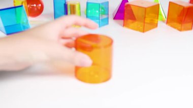 Eller, parlak bir ortamda eğitici ve oyuncu keşifleri vurgulayarak, beyaz bir yüzeyde canlı üç boyutlu geometrik şekilleri manipüle eder.
