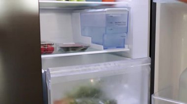 Parlak ışık altında çeşitli sebzeleri buzdolabı çekmecesine koyan eller, temiz ve düzenli bir mutfak ortamı sergiliyor.