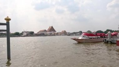Bir nehir teknesi Bangkok 'ta hareketli bir nehir boyunca ilerliyor, bulutlu gökyüzünün altında bir rıhtımdan geçiyor, yerel mimariyi gözler önüne seriyor.