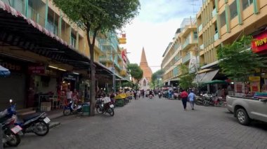 Yayalar ve motosikletler, gün ışığında sarı binalar ve meyve tezgahlarıyla dolu ve önünde bir tapınak kulesi bulunan canlı bir sokak pazarında geziniyorlar.
