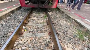 Bir yolcu treni, parlak gün ışığı ve yemyeşil ağaçların altında hareket eden insanlarla dolu bir açık hava pazar platformuna yavaşça yaklaşıyor. Statik, göz hizasında çekim