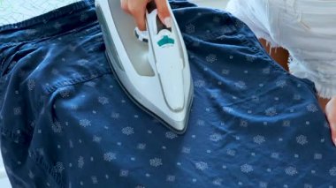 Bir kadın mavi desenli bir gömleği ütü masasına bastırmak için buhar ütüsü kullanıyor.