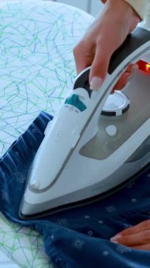 Bir insan mavi desenli bir gömleğin kırışıklıklarını düzeltmek için buhar ütüsü kullanır. Parlak, doğal ışık ve sabit kamera hareketleri.
