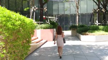 Bir kadın yeşil ve modern mimari ile çevrili güneşli bir şehir yolunda yürüyor. Sahne aydınlık ve huzurlu.