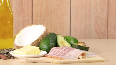 Avokado, hindistan cevizi, pastırma, tereyağı, fındık, yağ ve zeytinleri içeren çeşitli yüksek yağlı yiyecekler ahşap bir masaya dizilmiş, parlak stüdyo ışıklarıyla soldan sağa doğru pürüzsüz bir tavayla yakalanmış.