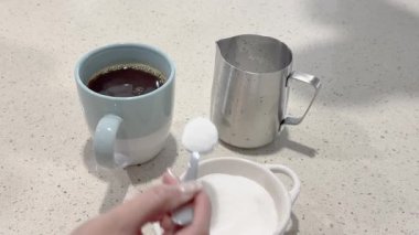 Bir el, mutfak tezgahındaki kahve fincanına şeker ekler ve sakin bir sabah atmosferi yaratır.