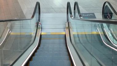 Sabit bir kamera temiz, boş bir yürüyen merdiveni yakalar. Parlak ışıklandırılmış, kartonla döşenmiş, cam parmaklıklı ve açık tabelalı bir alanda aşağı doğru hareket eder.